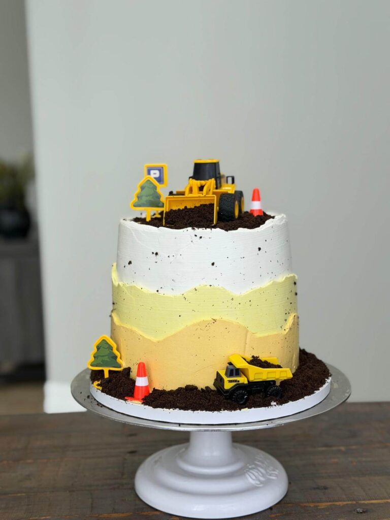 Construction-themed birthday cake with toy trucks, traffic cones, and textured buttercream details for kids’ celebrations.