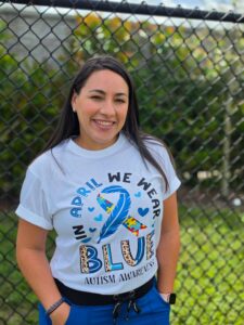 Smiling language therapist standing outdoors, wearing an autism awareness T-shirt featuring a blue ribbon and colorful puzzle pieces